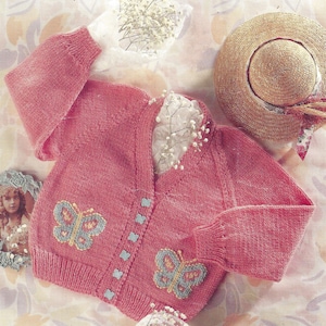 May include: A pink knitted cardigan with a V-neck and button closure. The cardigan has two butterfly designs on the front, one on each side of the buttons. The cardigan is laid out on a white surface with white flowers.