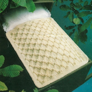 Könnte beinhalten: Eine cremefarbene gehäkelte Babydecke mit einem Rautenmuster. Die Decke liegt auf einem Kinderbett.
