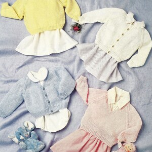 May include: Four knitted baby outfits in pastel colors are displayed on a blue surface. The outfits include a yellow sweater, a white cardigan, a light blue cardigan, and a pink sweater, each paired with a skirt or diaper cover. A stuffed animal and doll are also present.