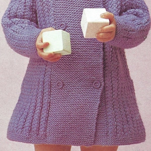 May include: A young child wearing a purple knitted cardigan with a button fastening. The child is holding two white blocks in their hands.