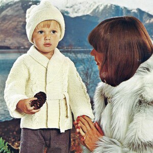 Peut inclure: Un enfant portant un cardigan en tricot crème avec un bonnet assorti. Le cardigan présente un motif en tricot torsadé et une ceinture. L'enfant tient une pomme de pin. L'arrière-plan montre un lac et des montagnes.