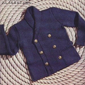 Könnte beinhalten: Ein dunkelblauer, zweireihiger Cardigan mit einem breiten Kragen und vier goldenen Knöpfen. Der Cardigan hat eine aufgesetzte Tasche auf der linken Seite und ist vor einem Hintergrund aus weißem Seil fotografiert.