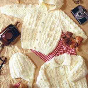 Puede incluir: Un conjunto de cardigan para bebé de crochet blanco con una chaqueta con capucha, un gorro a juego y cierres de botón. El conjunto está dispuesto sobre una superficie arenosa con osos de peluche y un paño de cuadros rojos y blancos.