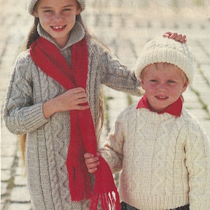 May include: Two children wearing matching cable knit sweaters and hats. The girl is wearing a red scarf. The boy is wearing a white sweater and a white hat.
