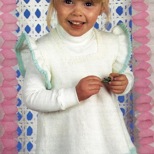 May include: A white knitted dress with a light blue trim and floral embroidery. The dress has a high neck and long sleeves. The girl is wearing the dress and has blonde hair.