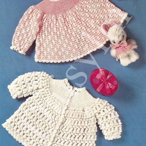 Könnte beinhalten: Zwei gehäkelte Baby-Pullover, einer rosa und einer weiß. Der rosa Pullover hat ein Spitzenmuster und der weiße Pullover hat ein strukturiertes, Noppenmuster. Beide Pullover haben einen gewellten Rand.