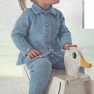 May include: A young child wearing a light blue knitted sweater and pants sits on a white toy duck. The duck has a yellow beak and black eyes.