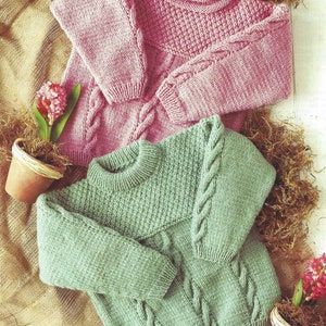 May include: Two knitted jumpers, one pink and one green, with a cable knit pattern. Both jumpers have a round neckline and long sleeves.