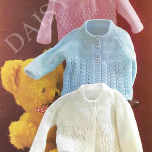 May include: Three knitted baby cardigans in pink, blue, and white with a button closure. The cardigans have a delicate lace pattern and are displayed on a wooden stand.