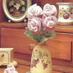 Puede incluir: Un ramo de rosas rosas de ganchillo en un jarrón con estampado floral. Las rosas están dispuestas en un racimo con hojas verdes de ganchillo. Hay una sola rosa rosa de ganchillo en la mesa junto al jarrón.