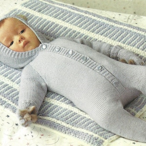 Peut inclure: Un bébé portant une combinaison en tricot à capuche bleu clair avec une fermeture à boutons. La combinaison a une capuche en forme d'ours avec des oreilles. Le bébé est allongé sur une couverture rayée blanche et bleue.