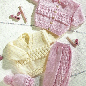 May include: A collection of hand-knitted baby clothes in soft pastel colors. The set includes a pink cardigan with heart-shaped buttons, a yellow sweater, pink pants, and a matching hat with a pom-pom. The garments feature textured patterns.