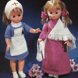 Op de afbeelding: Twee poppen gekleed in gebreide outfits. Eén pop draagt een blauwe jurk met een witte schort en een witte verpleegstersmuts met een rood kruis. De andere pop draagt een roze jurk met een witte sjaal en een witte gebreide tas.