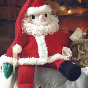 Pode incluir: Boneca de Papai Noel de tricô vestindo um terno vermelho com detalhes brancos, uma barba branca e um chapéu vermelho. Ela está segurando uma bolsa verde com uma etiqueta que diz "Feliz Natal".