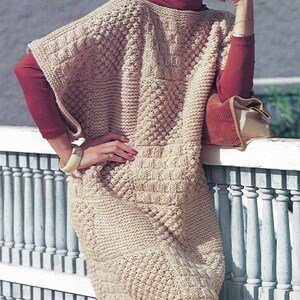 May include: A woman wearing a beige knitted poncho with a brown hat and red turtleneck sweater. She is standing by a white railing with a brown purse.