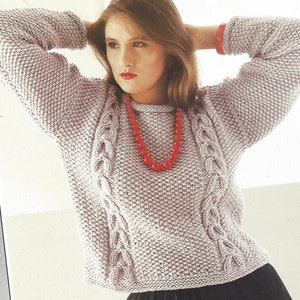 Puede incluir: Un suéter de punto gris claro con detalles de punto trenzado en la parte delantera. El suéter tiene cuello redondo y mangas largas. Se usa un collar de cuentas rojas. Las mangas tienen puños de tela roja.