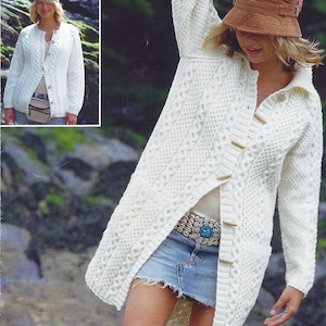 May include: A white knitted cardigan with a cable knit pattern and wooden buttons. The cardigan is worn over a white shirt and a denim skirt. The model is wearing a brown hat.