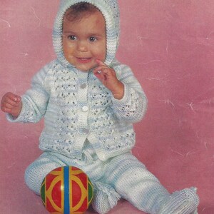 May include: A baby wearing a blue and white knitted sweater and pants set with a matching hat. The baby is sitting on a pink background and holding a red, yellow, and blue ball.