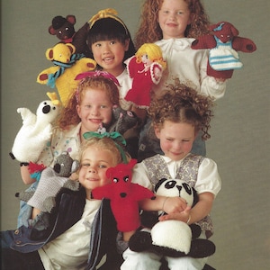 May include: A group of children are holding colorful knitted hand puppets. The puppets include a bear, a ghost, a koala, a red devil, a panda, a girl, and a dog. The children are smiling and looking at the camera.