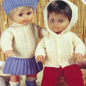 May include: Two knitted doll outfits, one with a blue beret and a white sweater and skirt, the other with a white hooded sweater and red pants.  The dolls are wearing the outfits and are standing on a sandy surface.