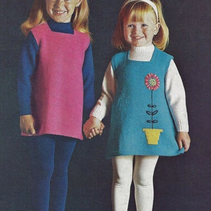May include: Two young girls wearing knitted dresses. The girl on the left is wearing a pink dress and blue tights. The girl on the right is wearing a blue dress with a flower design and white tights.