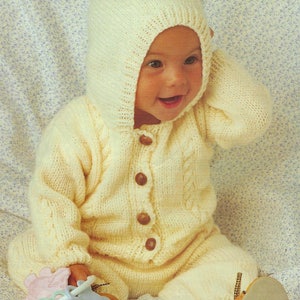 May include: A knitted baby outfit in a cream color with a hood and button closure. The outfit has a cable knit pattern and is worn by a baby sitting on a floral patterned surface.