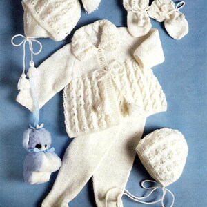 May include: A white knitted baby outfit set including a cardigan, pants, two hats, and a pair of booties. The set features a delicate cable knit pattern and is accented with white tassels.