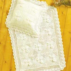 May include: A cream-colored, knitted baby blanket and pillow set. The blanket and pillow feature a floral pattern and a scalloped edge. The set is made of a soft, textured yarn, perfect for a nursery or as a decorative piece.