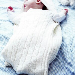 May include: A white knitted baby sleeping bag with a cable knit pattern. The sleeping bag has a hood and is made of soft, comfortable material.