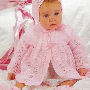 May include: A pink knitted baby outfit with a matching bonnet. The outfit features a delicate lace pattern and a bow at the neckline. The baby is wearing a floral print dress underneath.