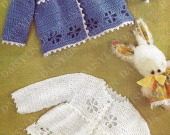 May include: Two crocheted baby jackets, one blue and one white, with a white crocheted trim and a floral design. The jackets are laid out on a green surface.
