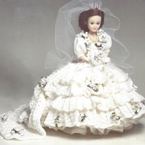 May include: A white crocheted wedding dress with a long train and a veil. The dress is decorated with white flowers and the doll is wearing a tiara.