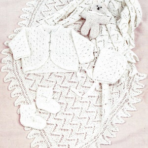 May include: A white crocheted baby set including a cardigan, bonnet, booties, and a blanket. The set is laid out on a pink surface with a small white crocheted teddy bear.