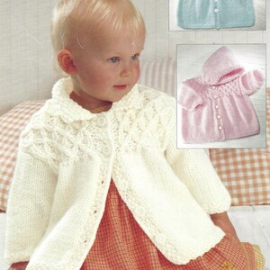 May include: A white knitted baby cardigan with a button closure and a delicate cable knit pattern. The cardigan is shown on a baby wearing a red and white gingham dress.