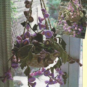 May include: A crocheted hanging flower basket with purple flowers and green leaves. The basket is hanging from a window and the flowers are cascading down.