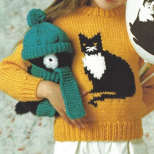 Pode incluir: Uma criança vestindo um suéter de tricô amarelo com um desenho de gato preto. O suéter tem um decote redondo e mangas compridas. A criança também está usando um chapéu e uma echarpe de tricô verde.