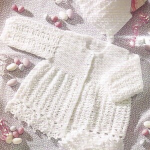 May include: A white crocheted baby outfit set, including a cardigan, a bonnet, and a diaper cover. The set is laid out on a floral patterned fabric with pink and white candy.