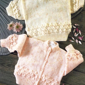 May include: Two knitted baby sweaters, one in a pale yellow with a speckled pattern and the other in a pale pink with a speckled pattern. Both sweaters have a textured design around the neckline and cuffs.