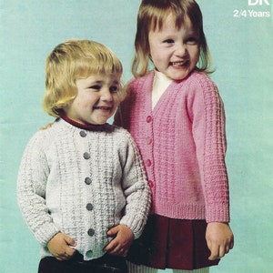 Puede incluir: Dos niños con cárdigans de punto. La niña de la izquierda lleva un cárdigan blanco con botones. La niña de la derecha lleva un cárdigan rosa con botones. Ambos cárdigans tienen un patrón de punto texturizado.