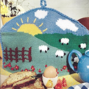Könnte beinhalten: Eine gestrickte Teewärmer mit einer ländlichen Szene mit blauem Himmel, gelber Sonne, weißen Wolken, grünen Feldern und Schafen. Ein brauner Zaun und rote Blumen sind ebenfalls zu sehen. Eine blaue Teekanne, ein Eierbecher und Toast sind im Vordergrund.