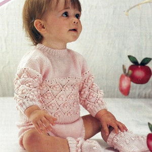 May include: A pink knitted baby sweater and matching socks with a lacy pattern and bobbles. The sweater has a round neckline and long sleeves.
