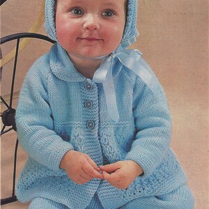 May include: A baby wearing a light blue knitted cardigan and matching trousers with a light blue ribbon tied around the neck. The cardigan has a button fastening and the trousers have a wide leg.