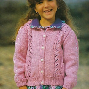 May include: A pink knitted cardigan with a button closure and a lace pattern. The cardigan has a purple collar and is worn over a floral print dress.