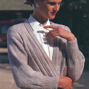 May include: A light purple knitted cardigan with a V-neck and button closure. The cardigan has a cable knit pattern and is worn over a white button-down shirt.