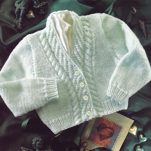 May include: A light blue knitted baby cardigan with a cable knit design on the front and sleeves. The cardigan has a shawl collar and is fastened with four white flower-shaped buttons. The garment is displayed on a dark green satin surface.