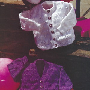 Puede incluir: Dos cárdigans de punto para bebés. Uno es blanco con un patrón de corazón y el otro es morado con un patrón de corazón. Ambos cárdigans tienen botones en la parte delantera.