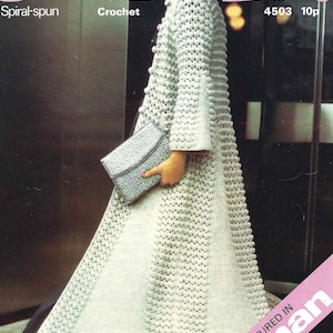 May include: A white crocheted coat with a textured pattern. The coat has a high collar and long sleeves. The model is wearing a black hat and holding a silver clutch.