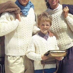 Könnte beinhalten: Eine dreiköpfige Familie trägt weiße Strickpullover mit Zopfmuster. Die Pullover haben ein traditionelles irisches Design. Der Mann trägt einen blauen Schal und der Junge trägt eine braune Ledertasche.