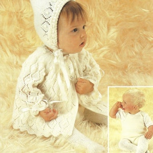 May include: A white knitted baby outfit with a matching bonnet. The outfit features a lacy design and a button closure. The baby is wearing the outfit and sitting on a fluffy white surface.