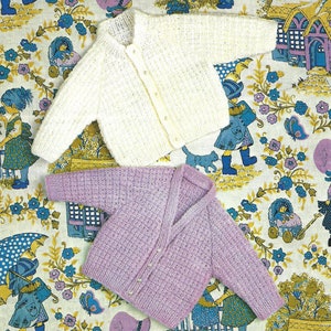 Puede incluir: Dos cárdigans de punto para bebé, uno blanco y otro morado, con cierres de botón. Los cárdigans están colocados sobre un fondo de tela con estampado floral.